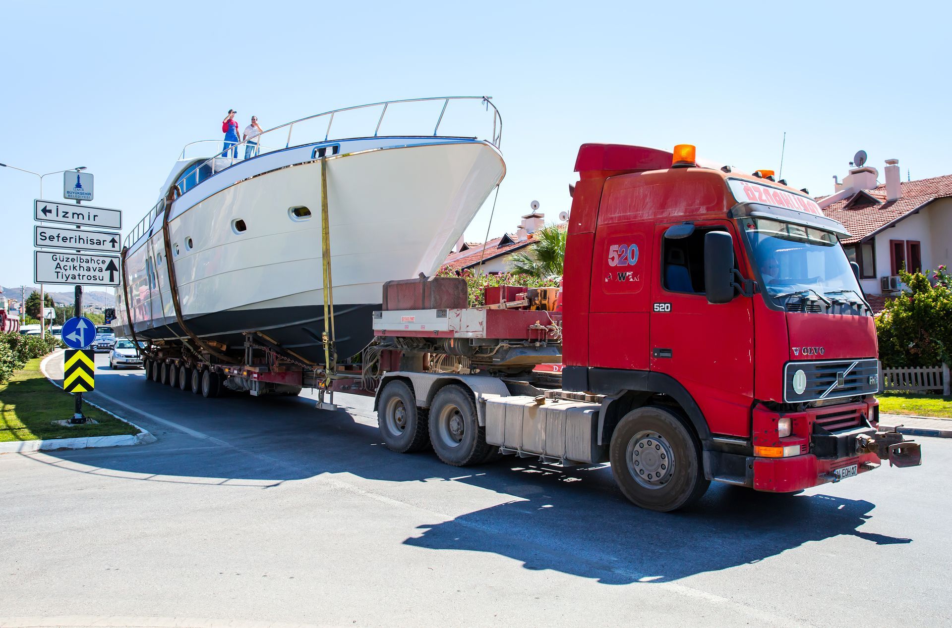 A large red truck transporting a white yacht on a trailer. People stand on the yacht.