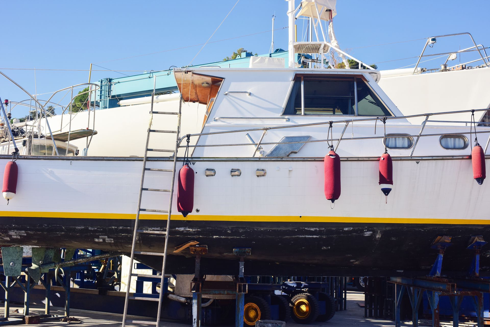 White boat on blocks, with a ladder and red fenders. Yellow trim, blue sky.