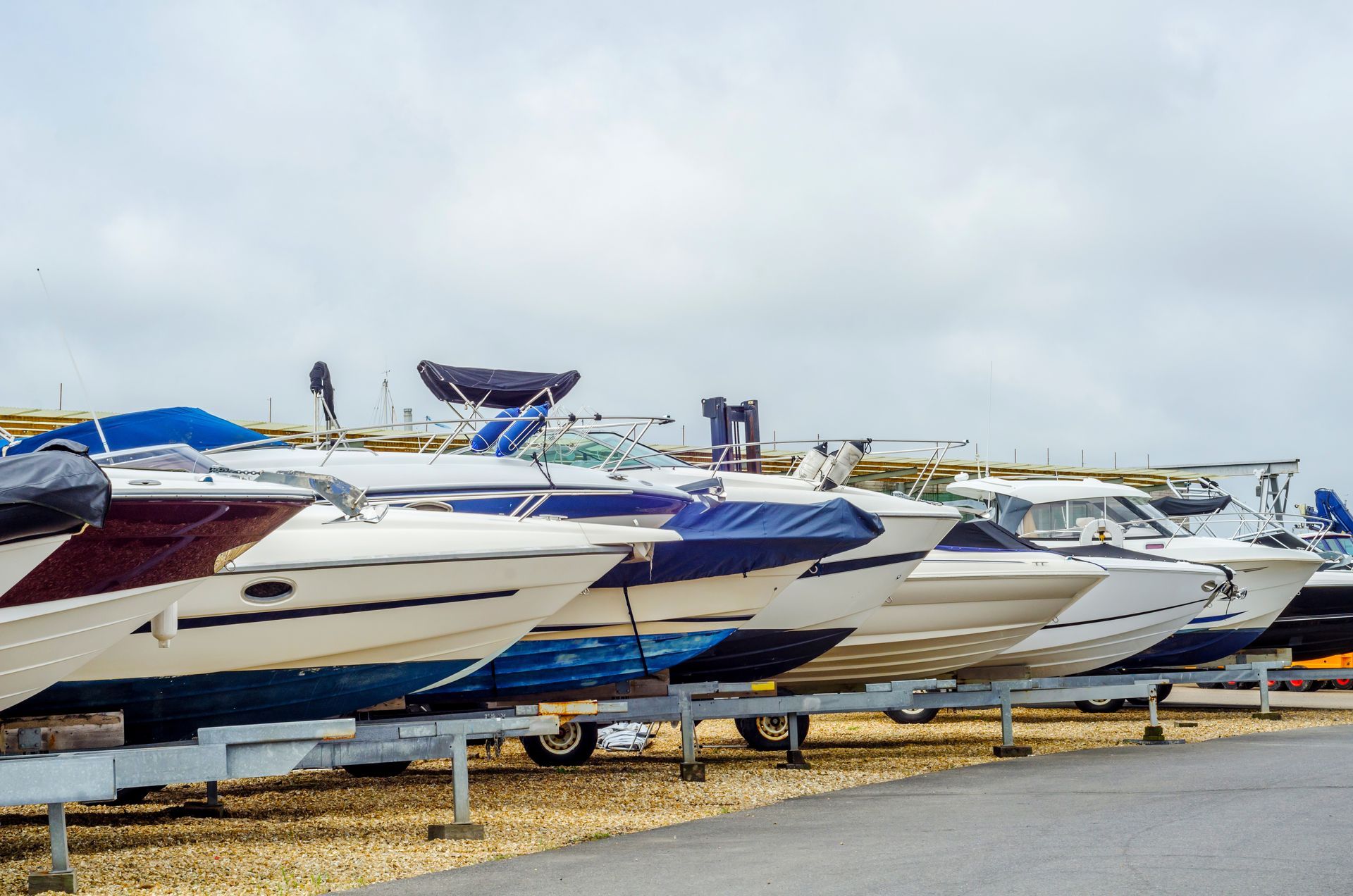 Boats covered in blue and burgundy tarps on trailers, parked along a paved area under a cloudy sky.