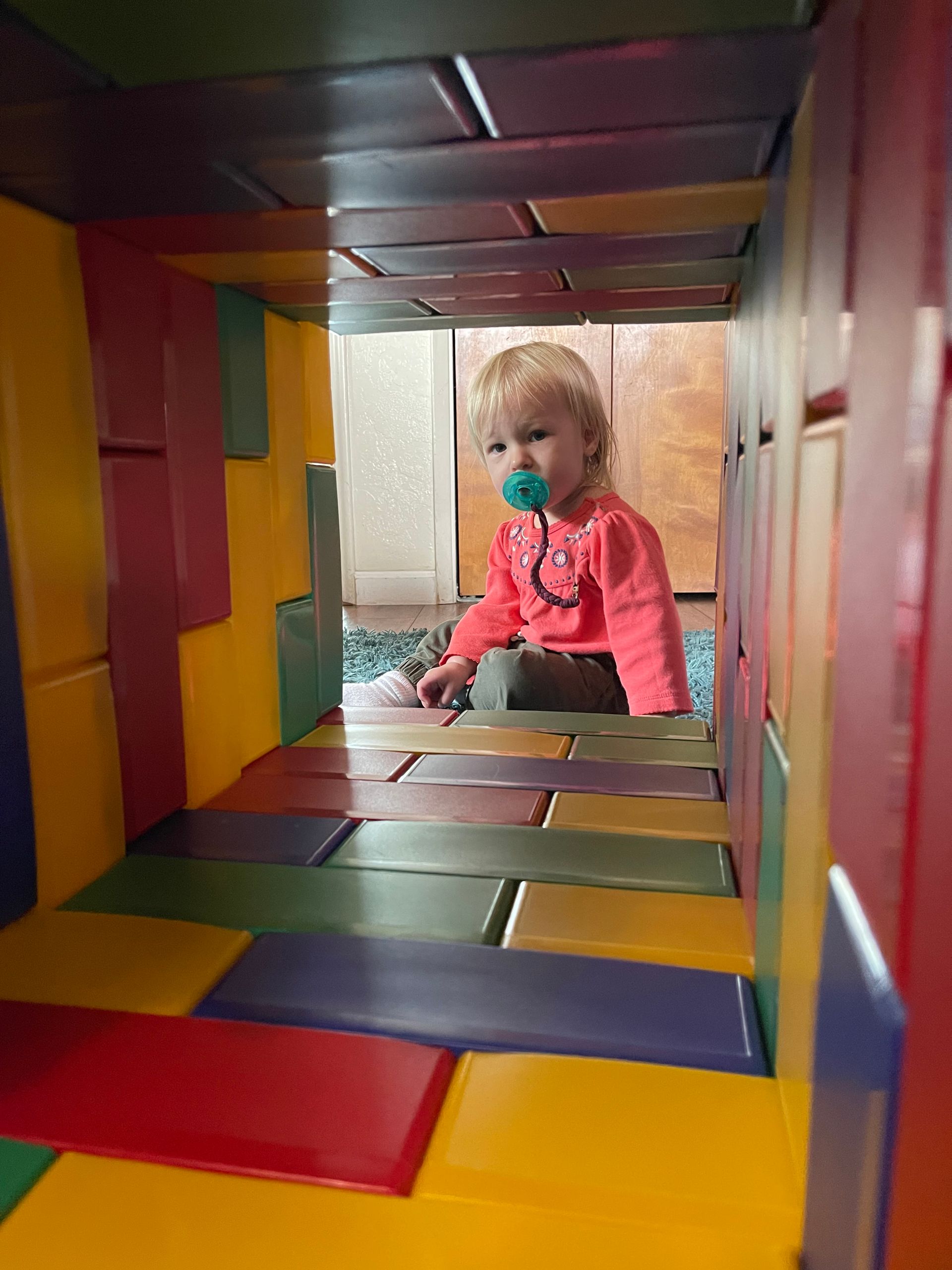 Child inside a colorful play structure, looking forward with a pacifier.