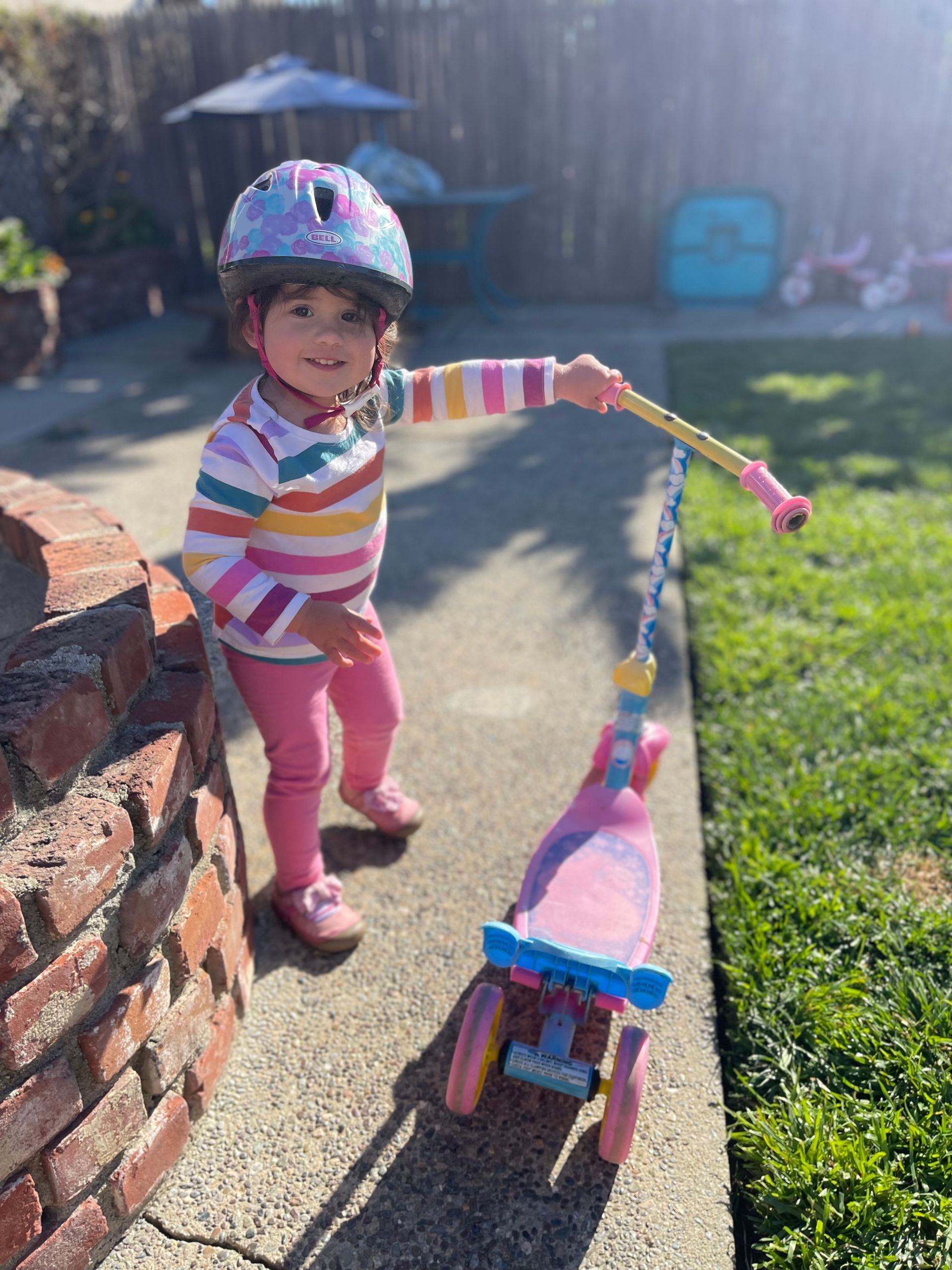 Child in helmet and colorful striped shirt holding a pink scooter on a sunny patio.