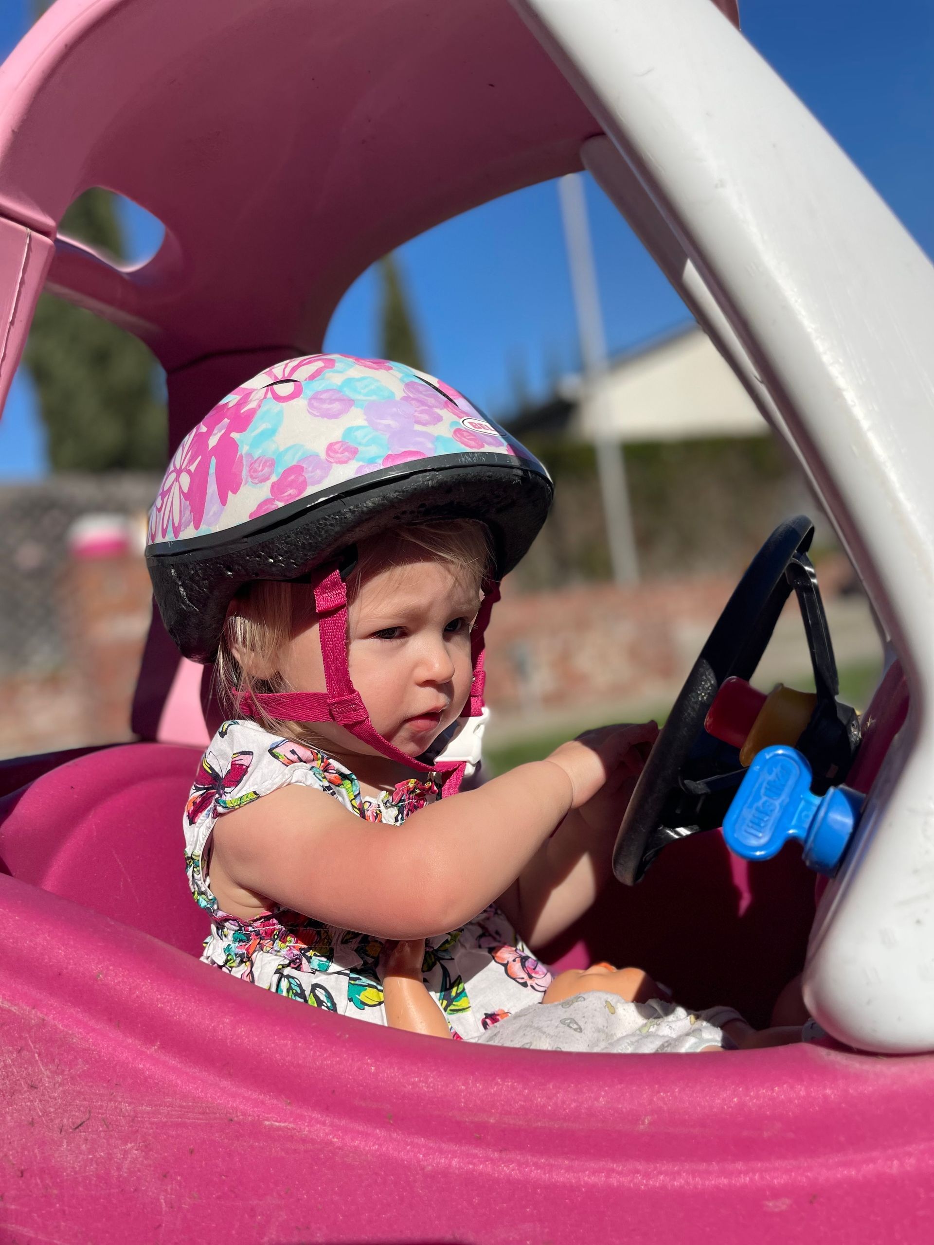 Toddler in a pink toy car wearing a floral helmet, hands on steering wheel, outdoors.
