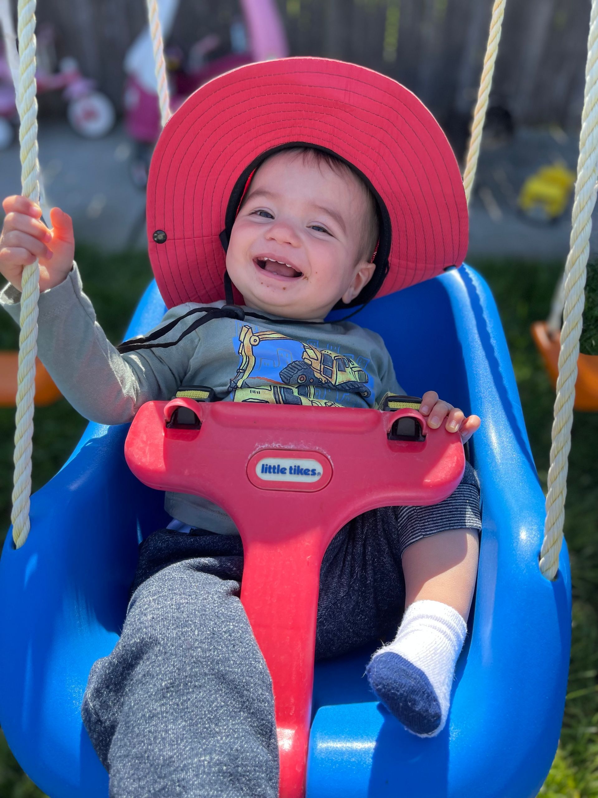 Smiling child in a blue swing with a red hat, holding onto the swing's safety bar, in a sunny outdoor setting.