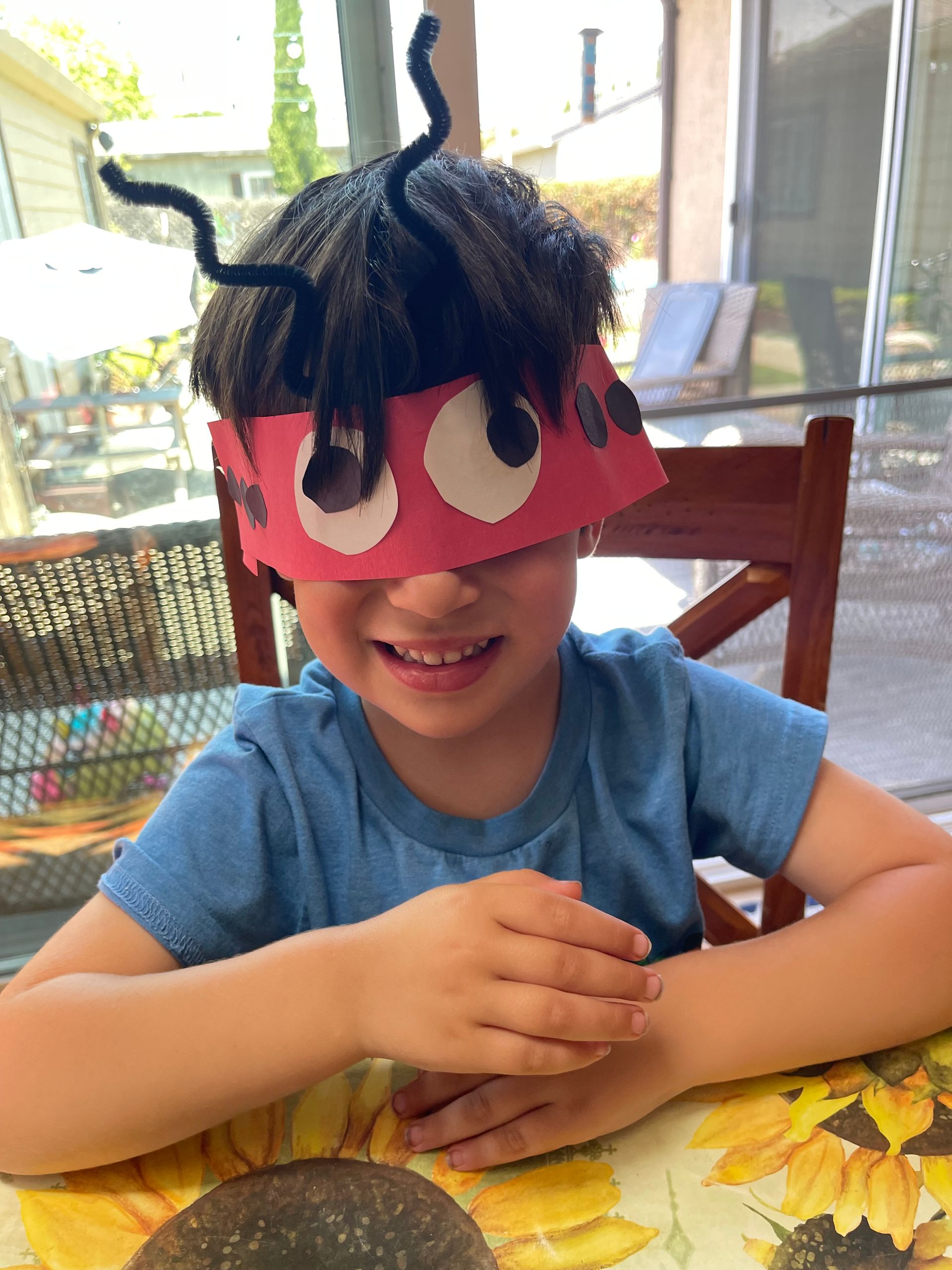 A young boy wearing a paper ant headband is sitting at a table.