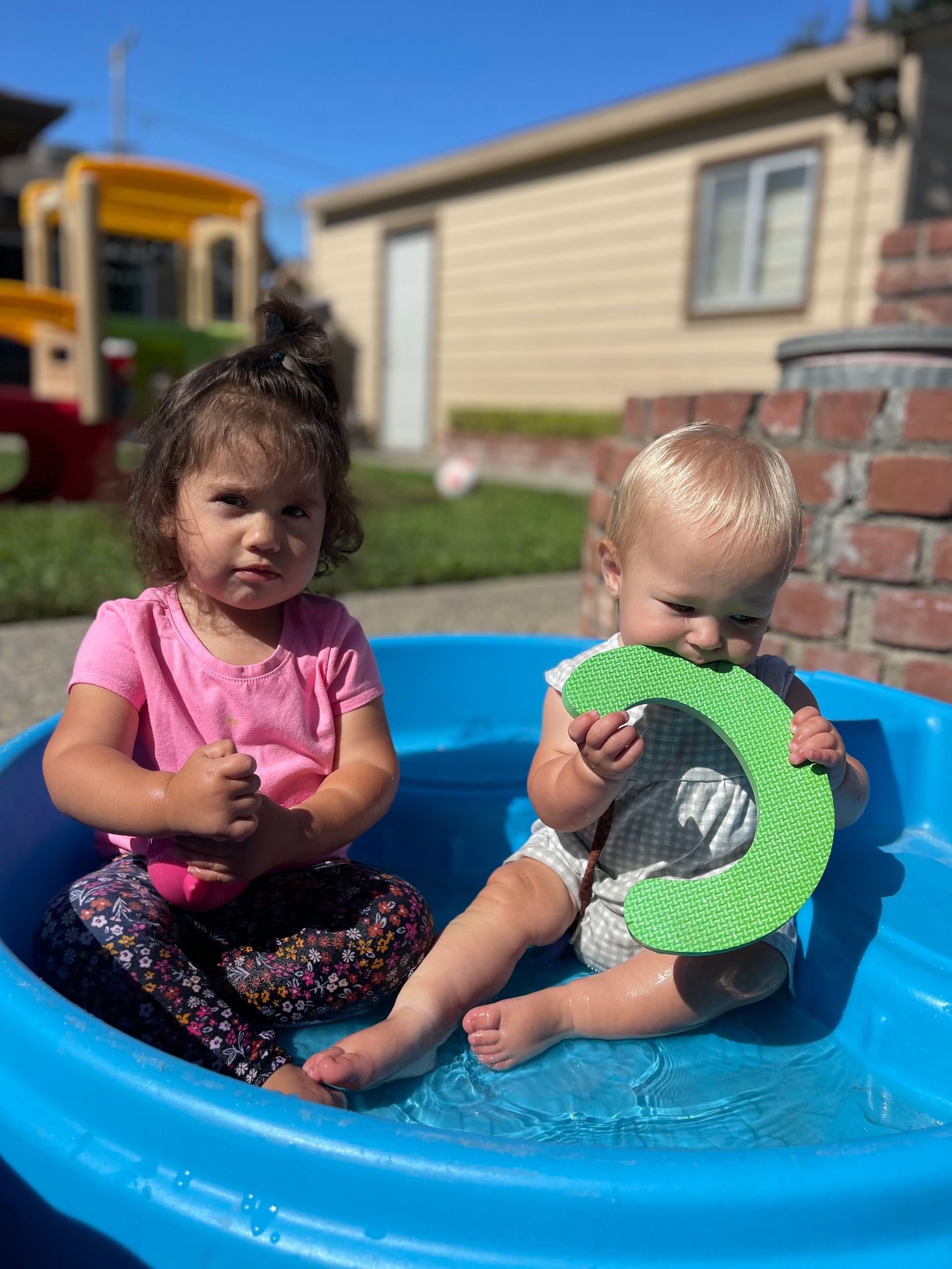 Two children in a blue plastic pool. One chews a green