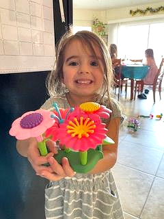 Girl smiling, holding pink and yellow toy flowers; a dining table and others in background.