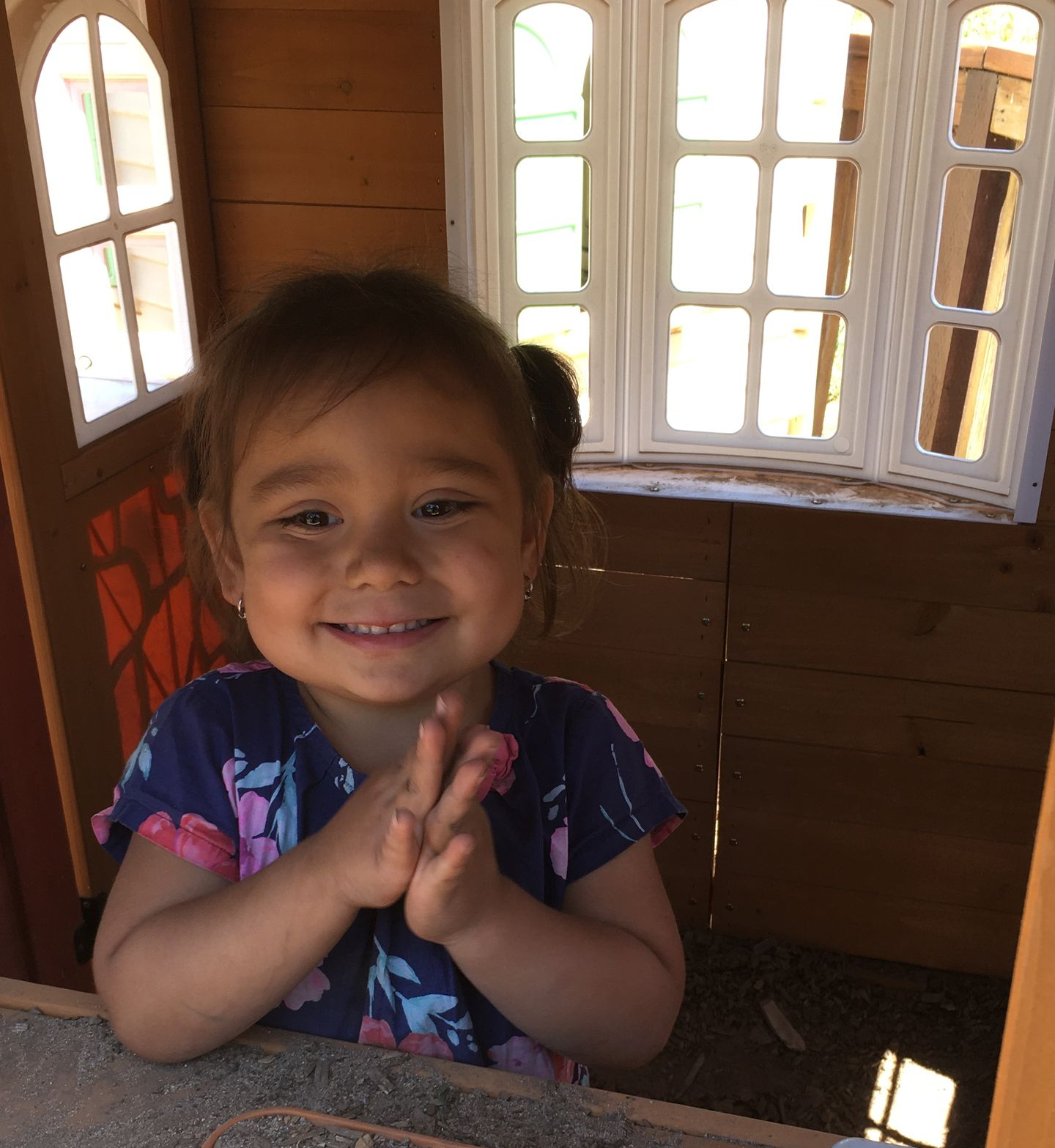 Girl smiling inside a wooden playhouse with hands clasped. Bright sunlight illuminates windows.