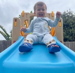 Toddler smiles on a blue slide. Wearing blue shoes and a light blue onesie. Outdoors.