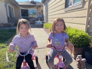 Two young children riding pink tricycles on a path. Smiling, outdoors, sunny day.