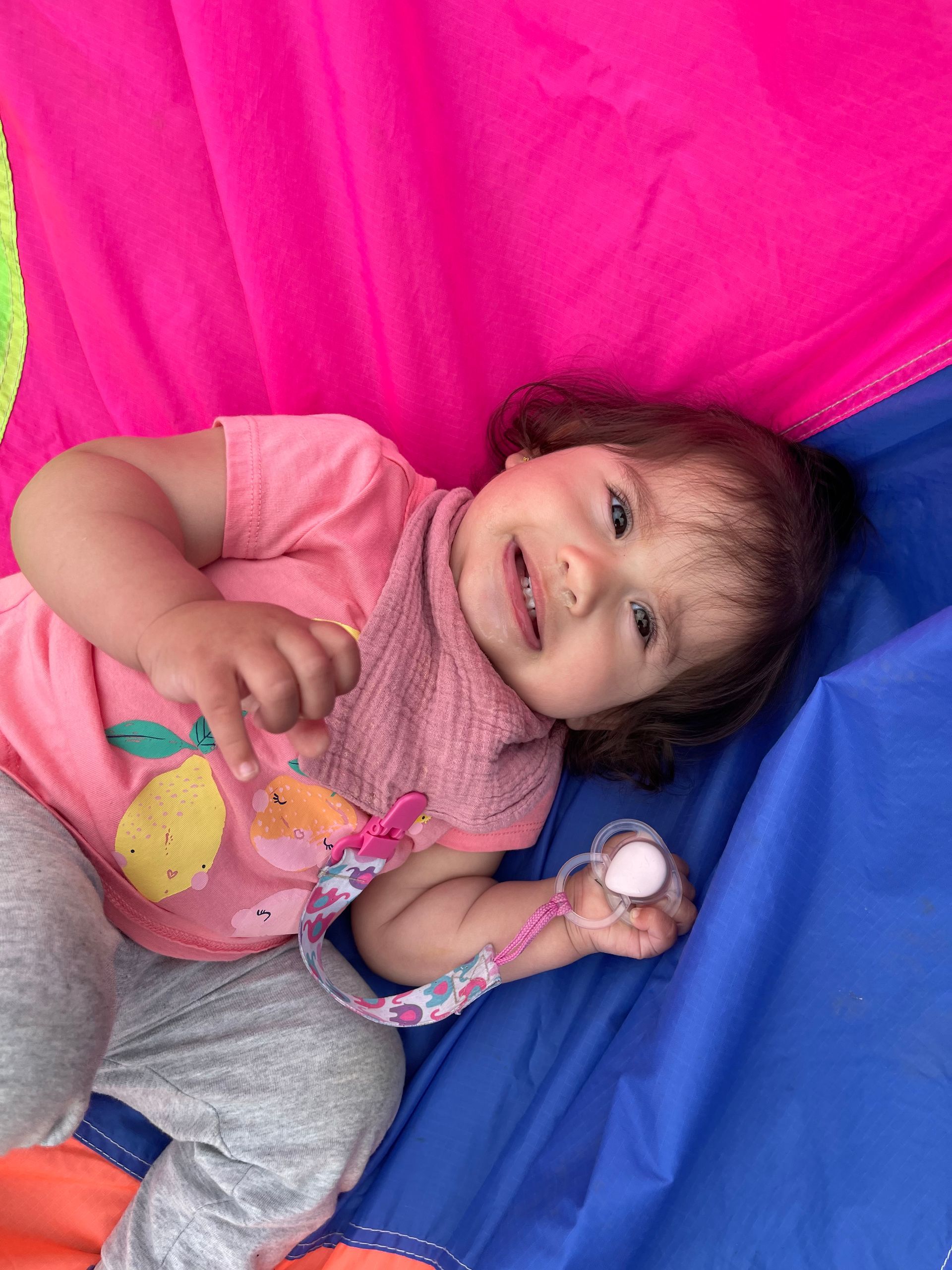 Child lying on back, smiling, with a pacifier, wearing a pink shirt, set against a pink and blue background.