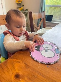 A baby in a high chair plays with a pink owl-shaped sewing card on a wooden table.