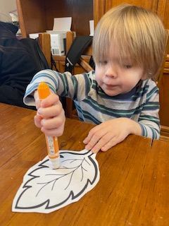 Young child coloring a leaf-shaped outline with an orange marker at a table.