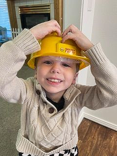 A young child in a yellow construction helmet smiles, adjusting the helmet with both hands.