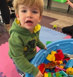 Young child in green hoodie holding a blue bin full of colorful blocks.