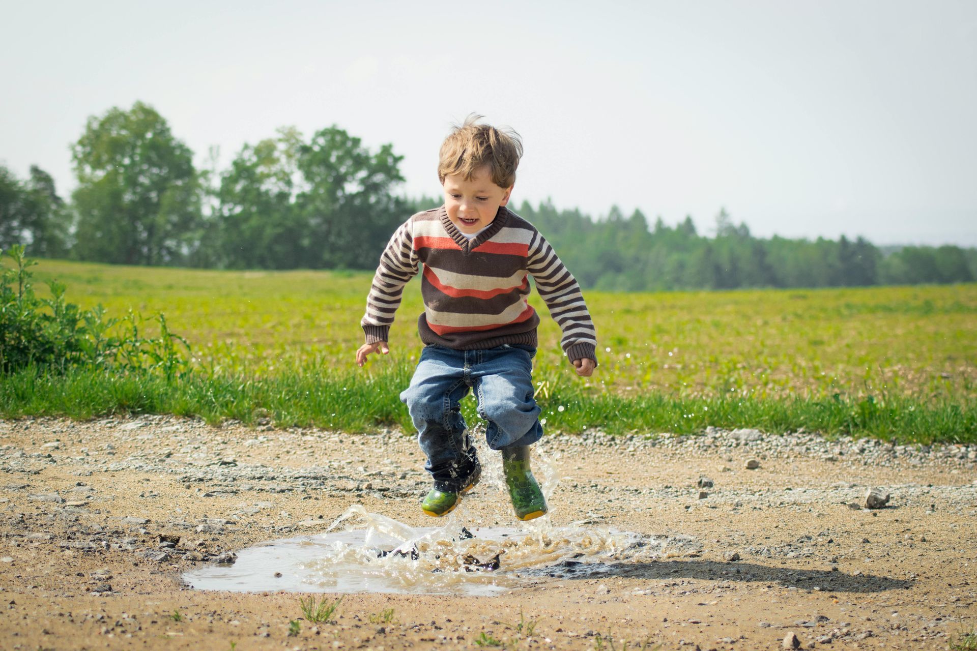 Boy in striped shirt jumps into a puddle on a dirt path in a field, smiling.