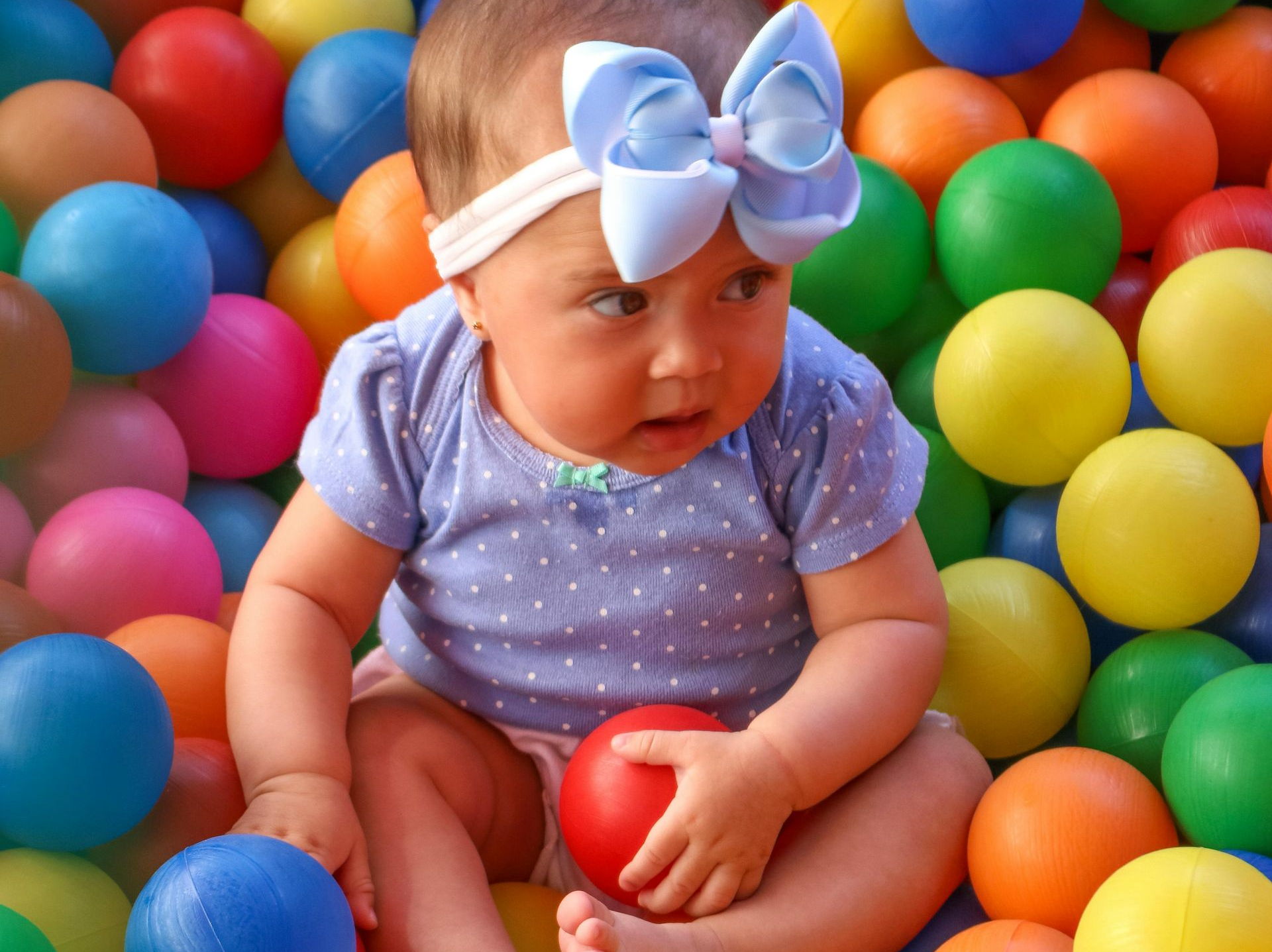 Baby in a ball pit, wearing a light blue bow and a dotted shirt, holding a red ball.