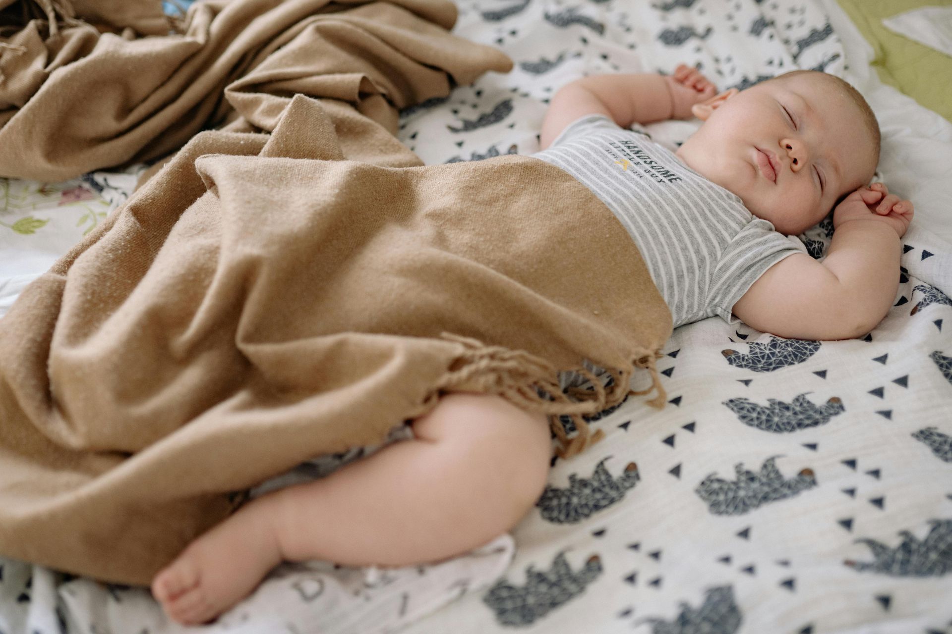 Sleeping baby under a tan blanket on a bed with a patterned sheet.