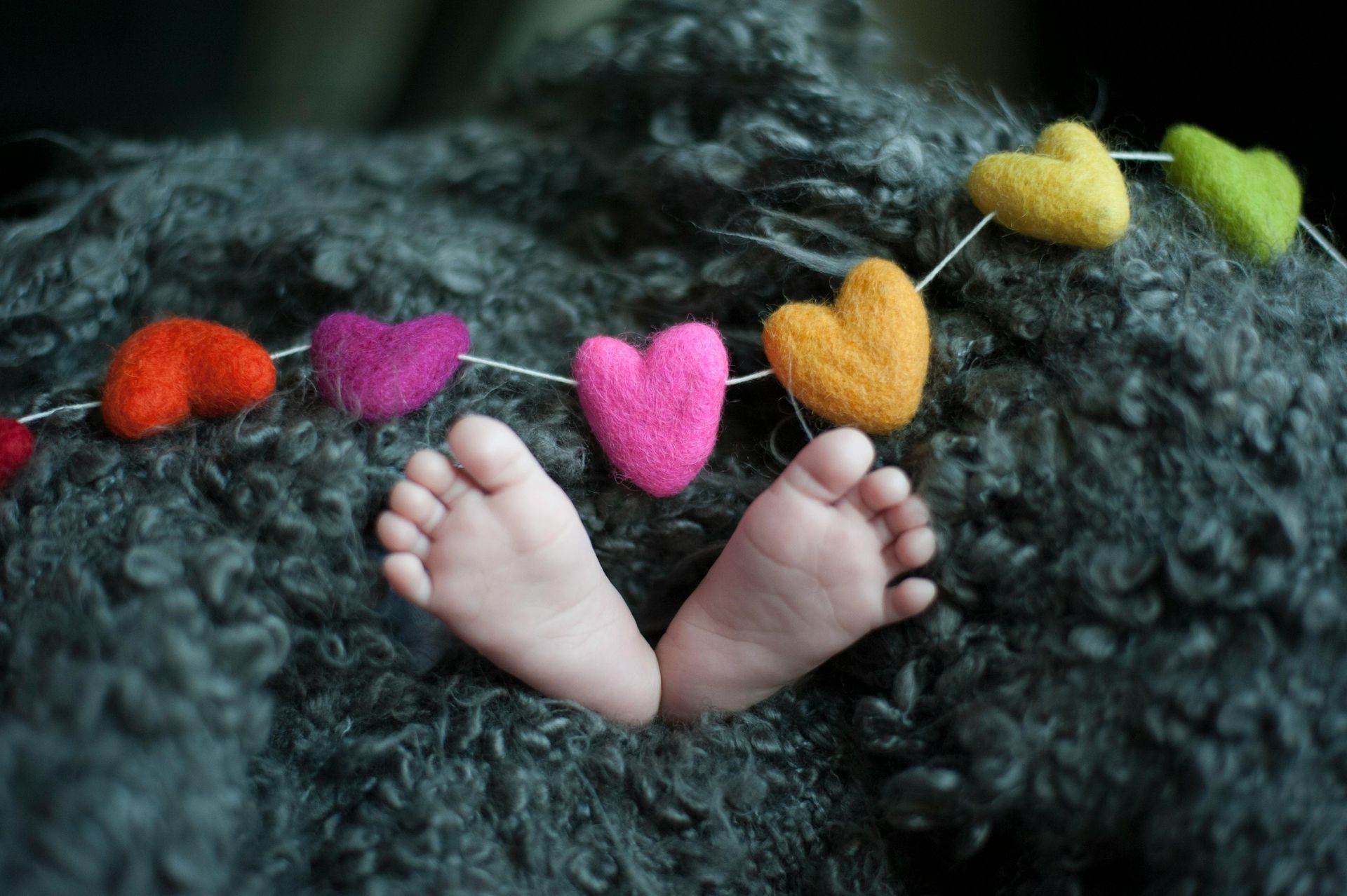 Baby feet in front of a garland of colorful felt hearts on a fuzzy gray blanket.