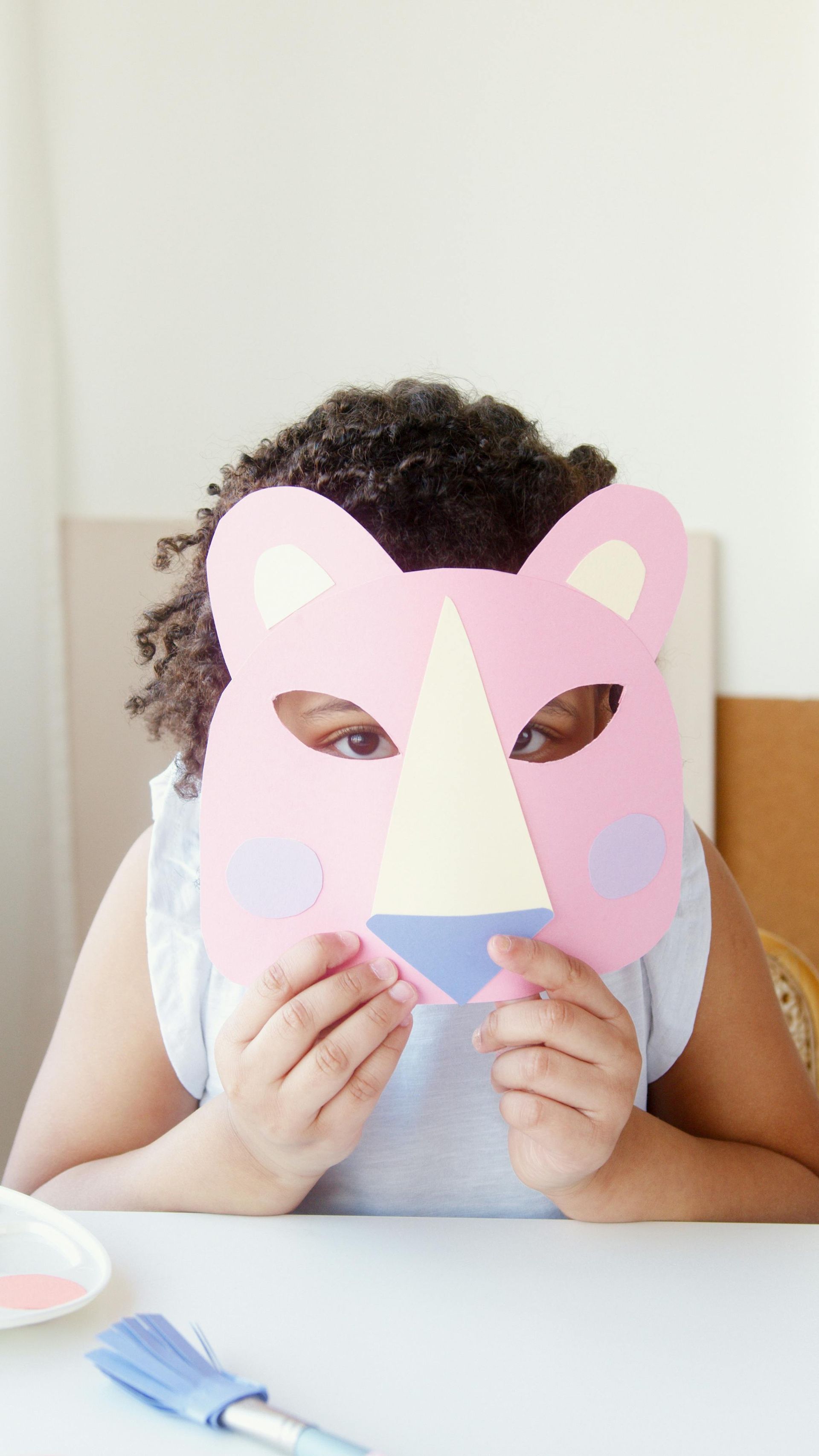Girl holding a pink paper animal mask in front of her face; craft supplies visible.