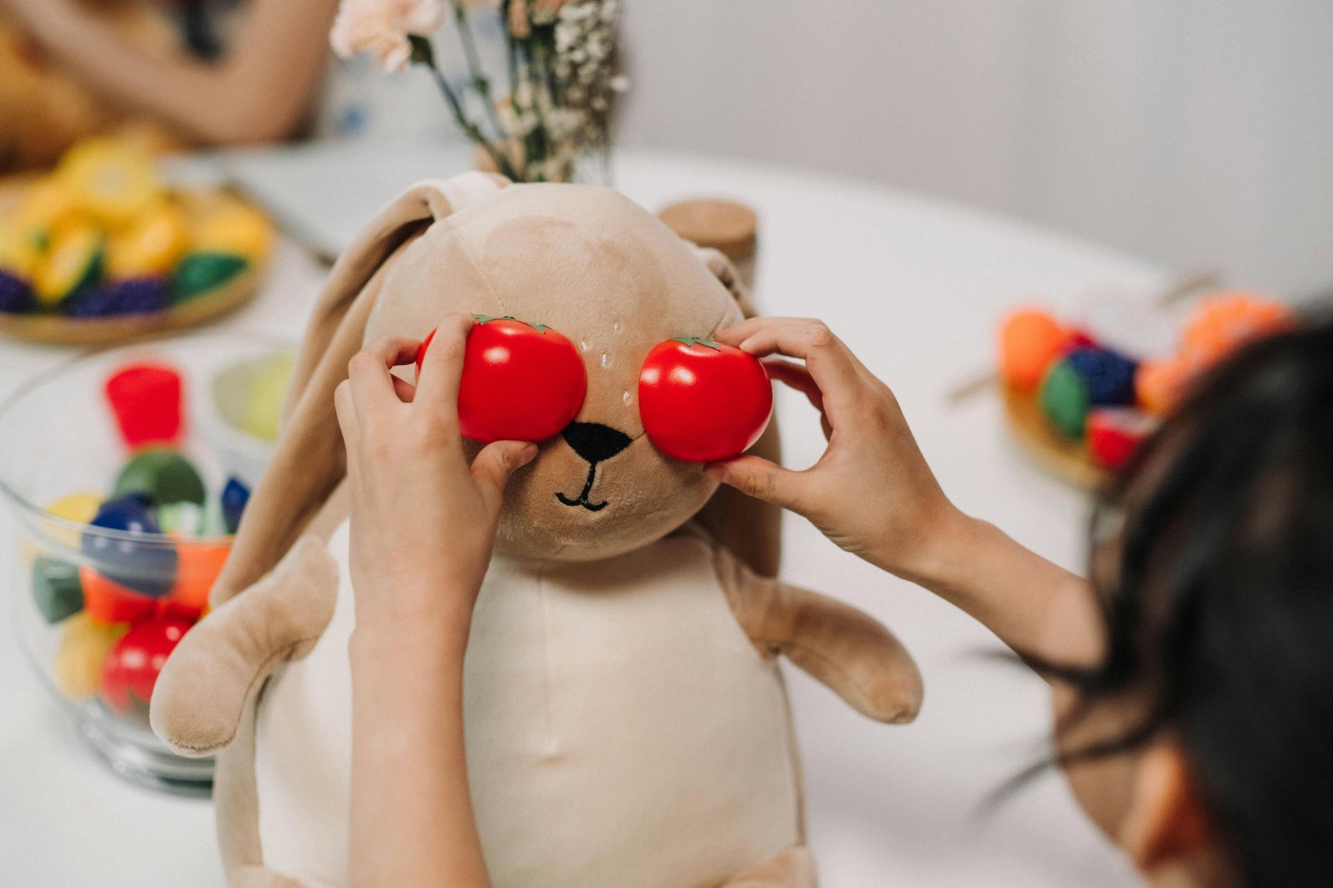A child placing two red tomatoes on a stuffed bunny's eyes at a table.