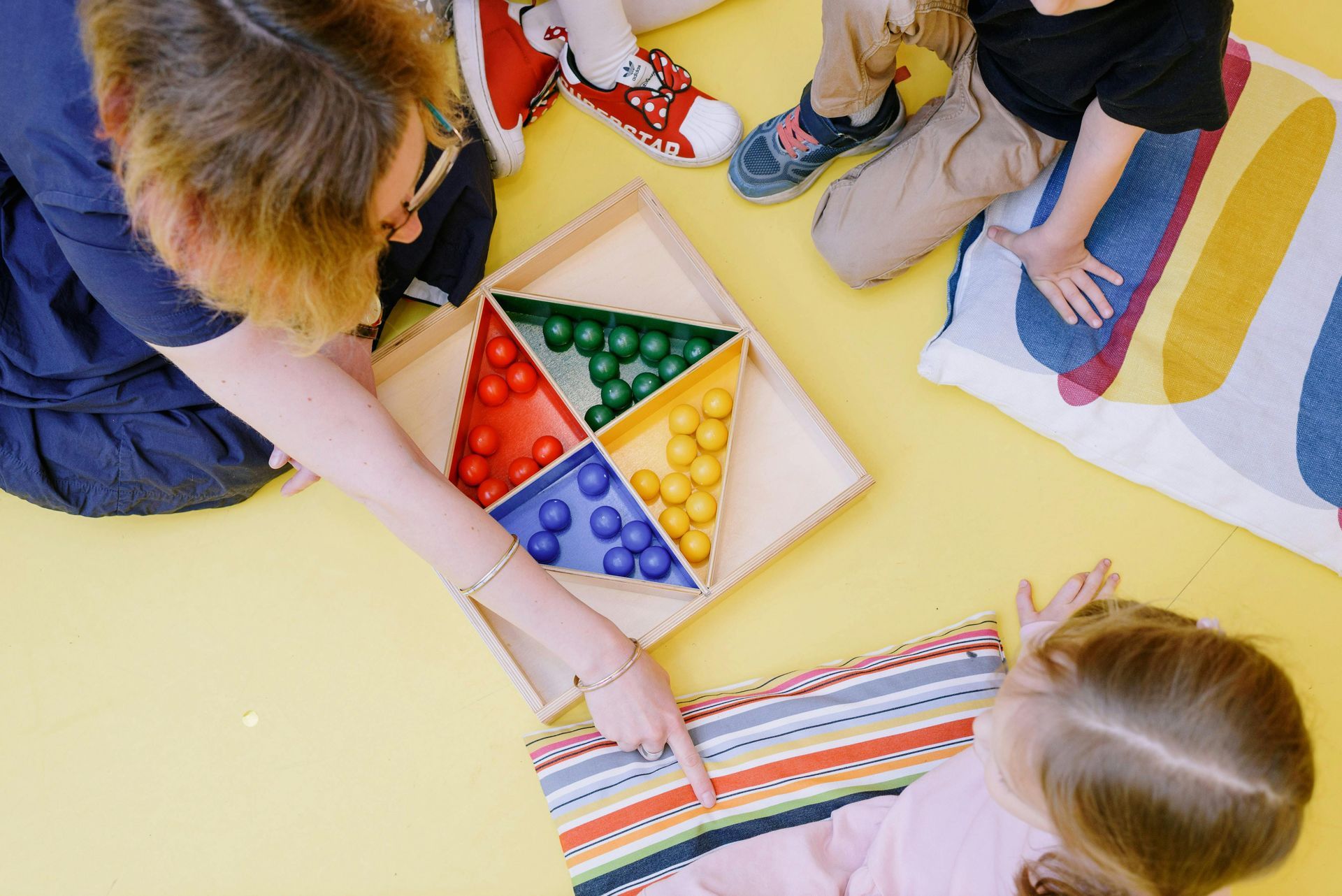 Teacher points to striped fabric while children play with a wooden toy with colorful balls on a yellow floor.