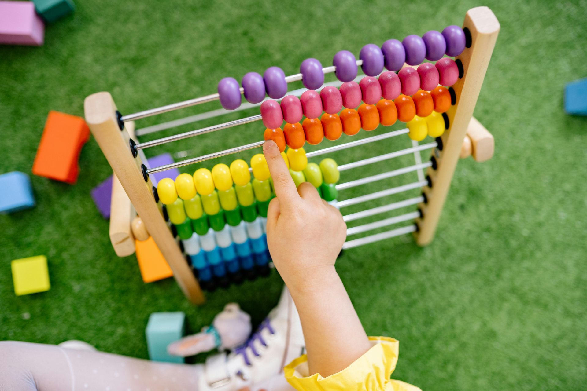 Child's hand pointing at orange beads on a colorful abacus. Green turf background with toys.