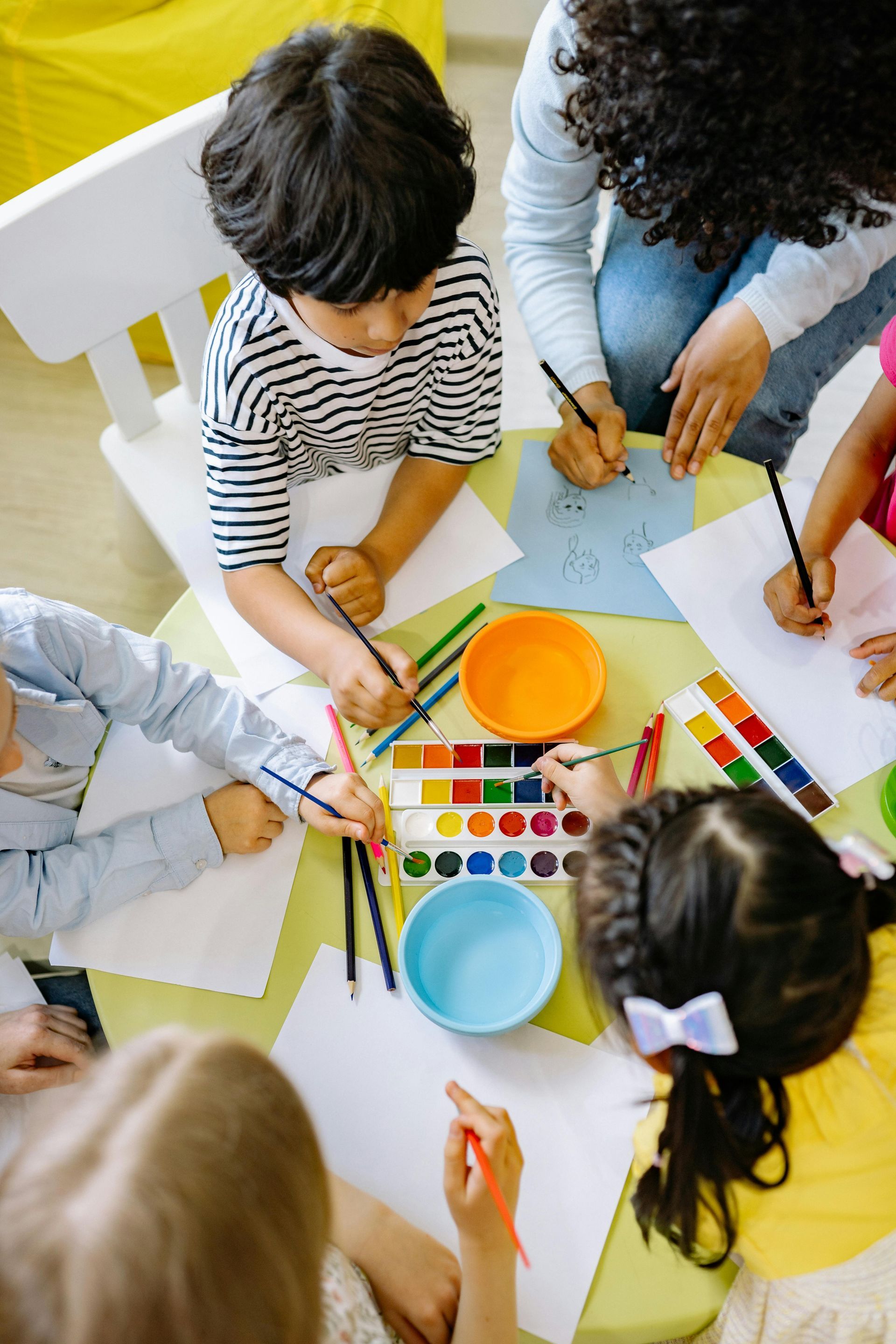 Children and adult drawing at a table, using paint and pencils.