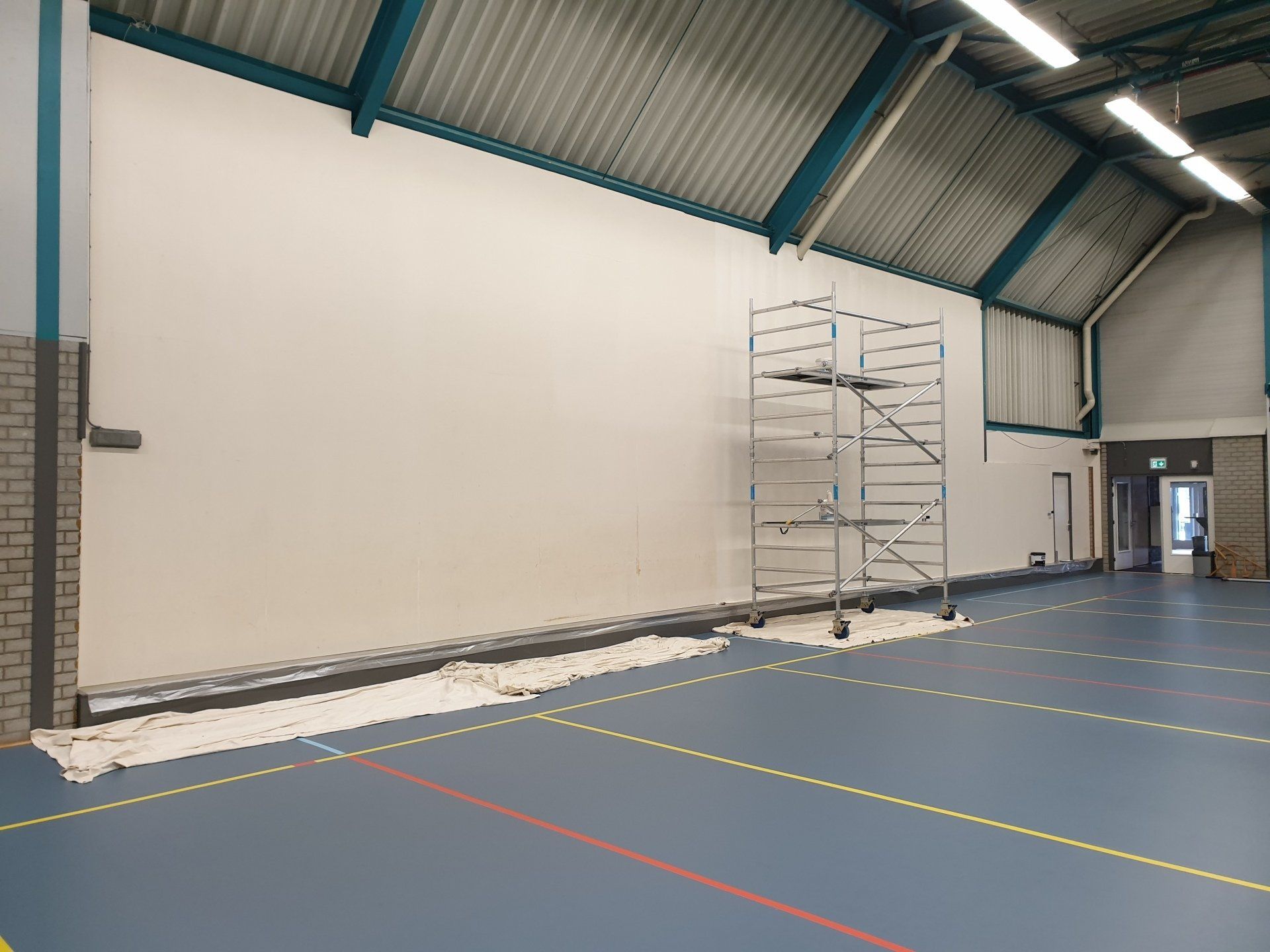 Interieur van een sportschool met steigers bij een grote witte muur, schilder werkzaamheden in uitvoering.