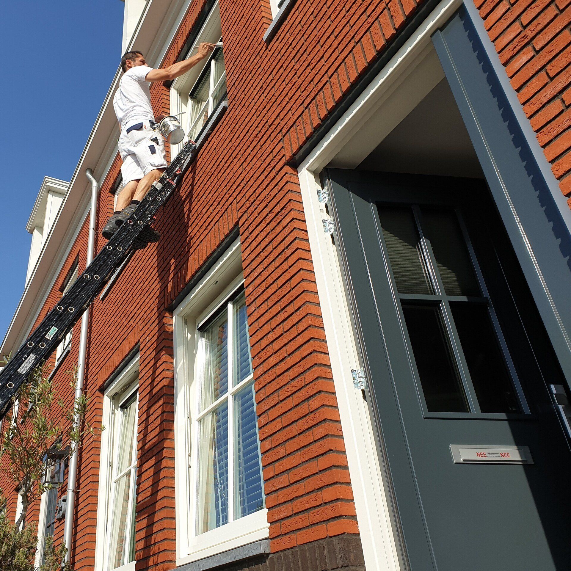 Stefan op een ladder schildert een raam in een bakstenen gebouw met een Antracietgrijze deur op een zonnige dag.
