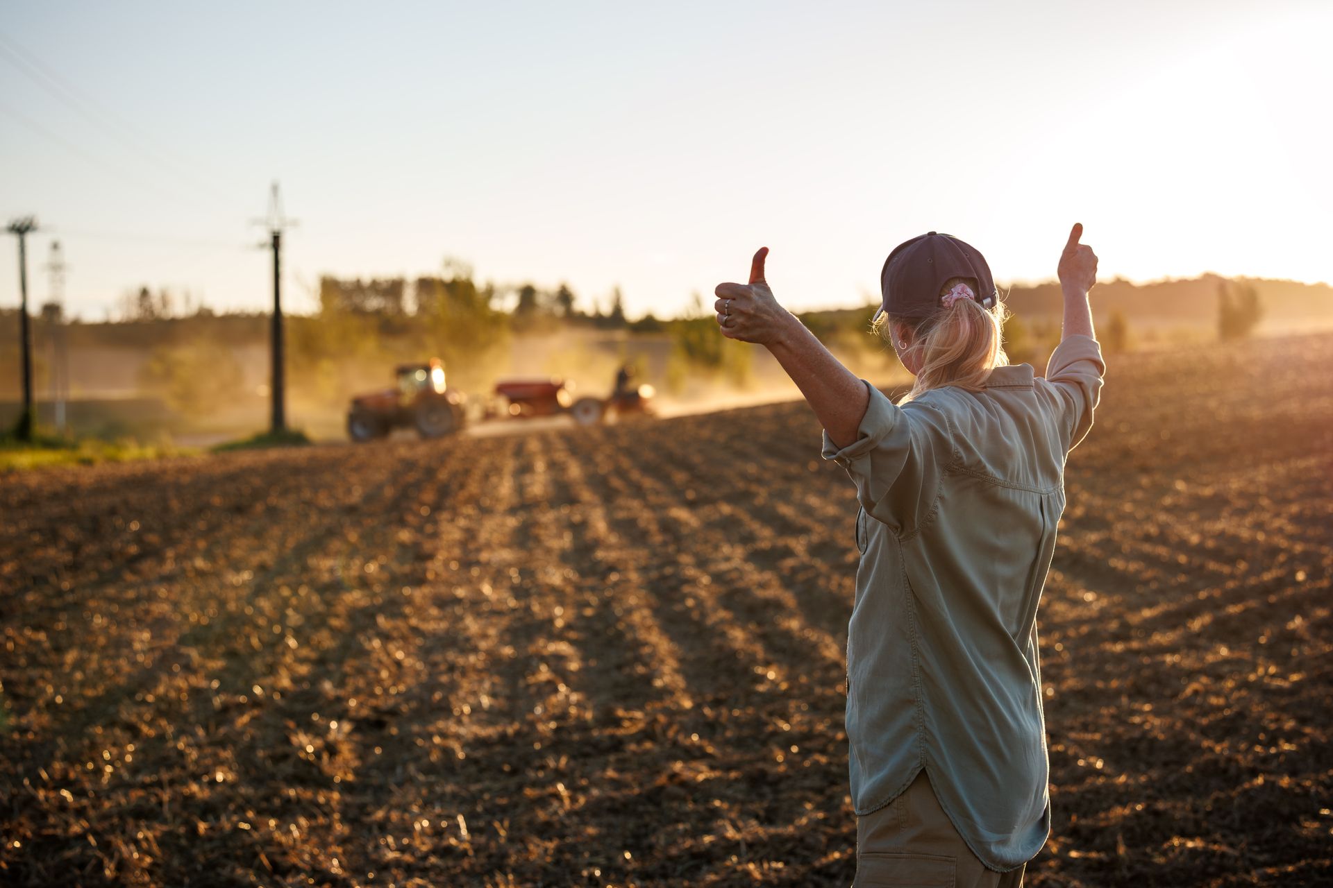 A farmer raising her arms with thumbs up after a sowing process by agricultural machinery.