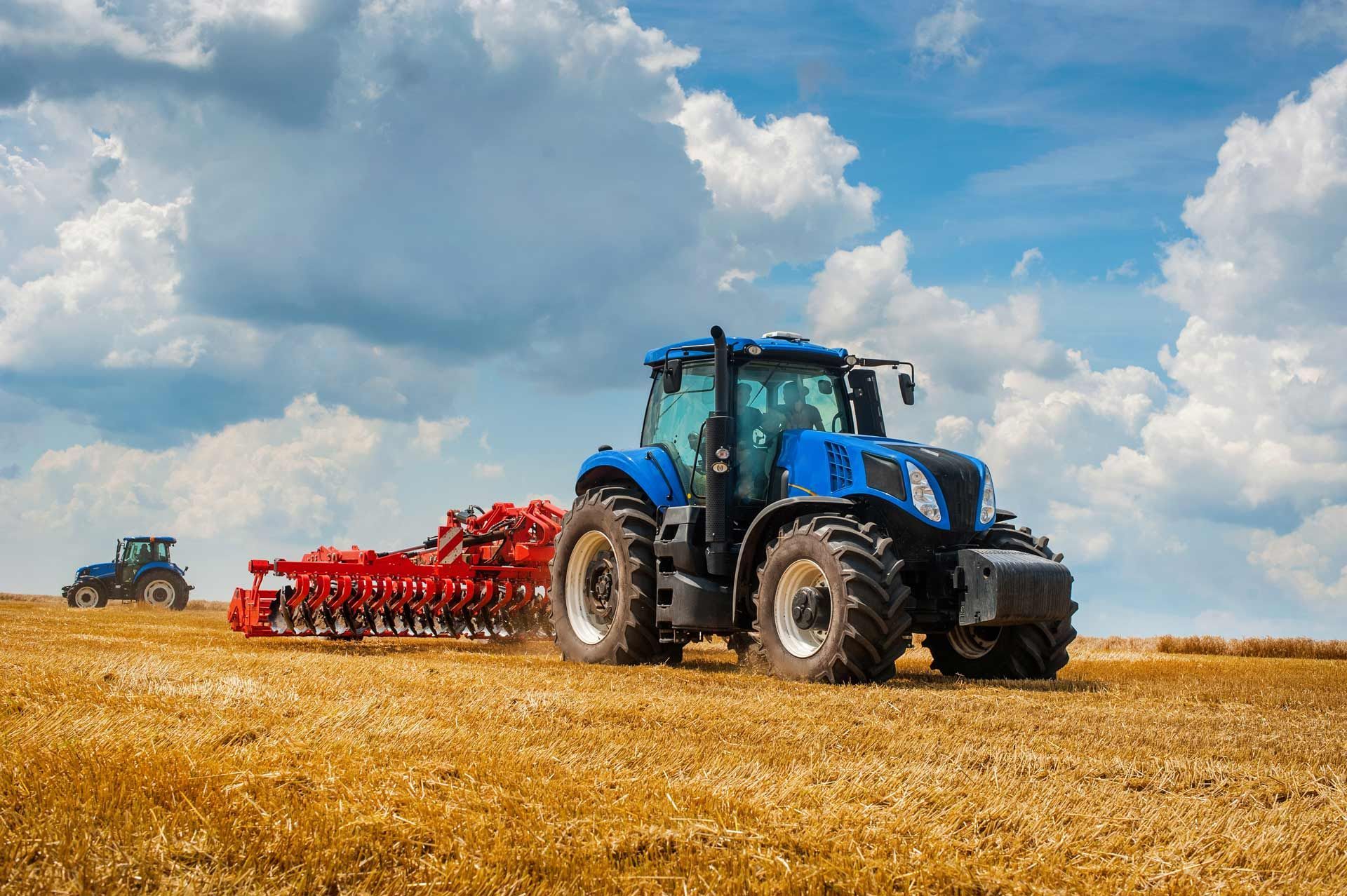 A new blue tractor with a red harrow in a field against a cloudy sky.