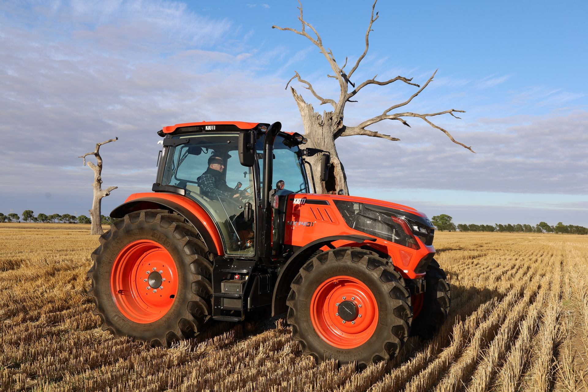 Orange tractor in a field, under a blue sky, with a person driving. Dead trees in the background.