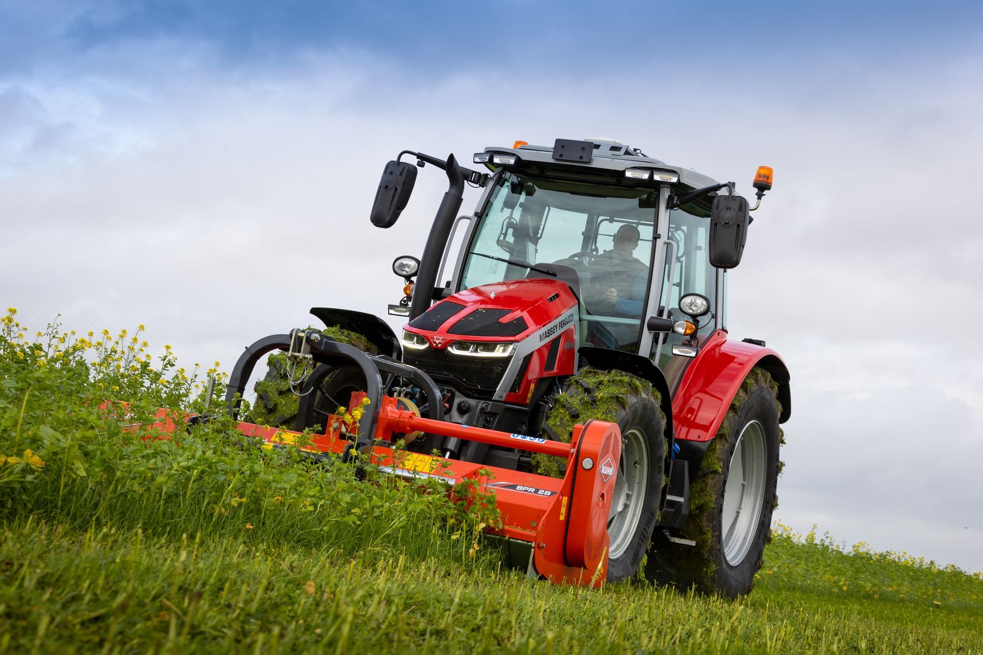 Red tractor mowing tall grass in a field under a cloudy sky.