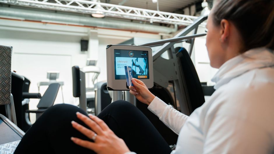 Een vrouw zit op een apparaat in een sportschool en gebruikt een tablet.