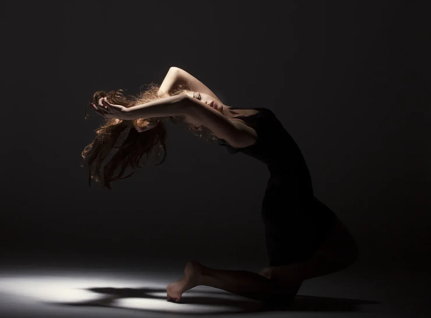 Woman in a black dress arches backward, illuminated by a spotlight on a dark background.