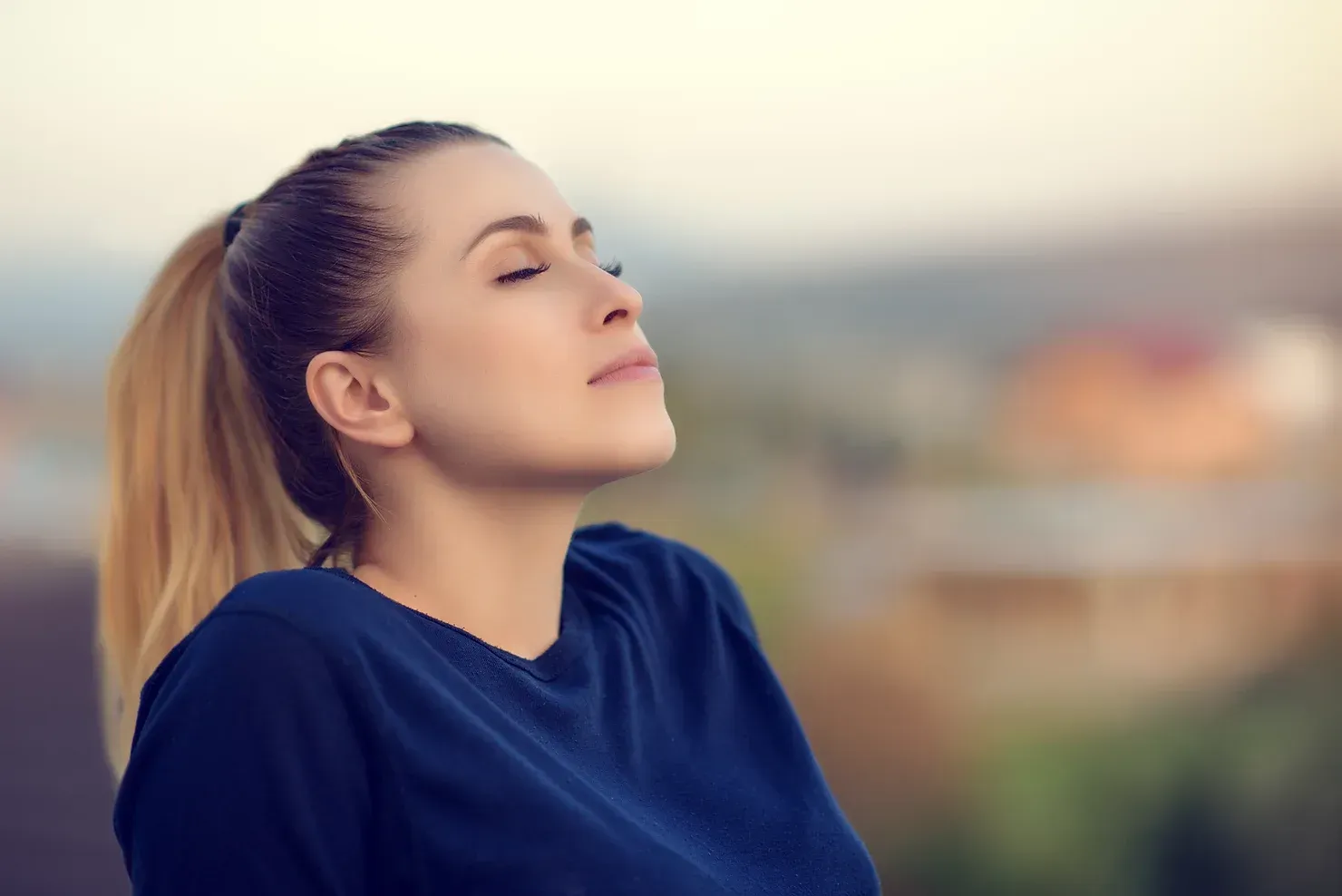 Woman with blonde ponytail, eyes closed, and head tilted up, taking a deep breath outdoors.
