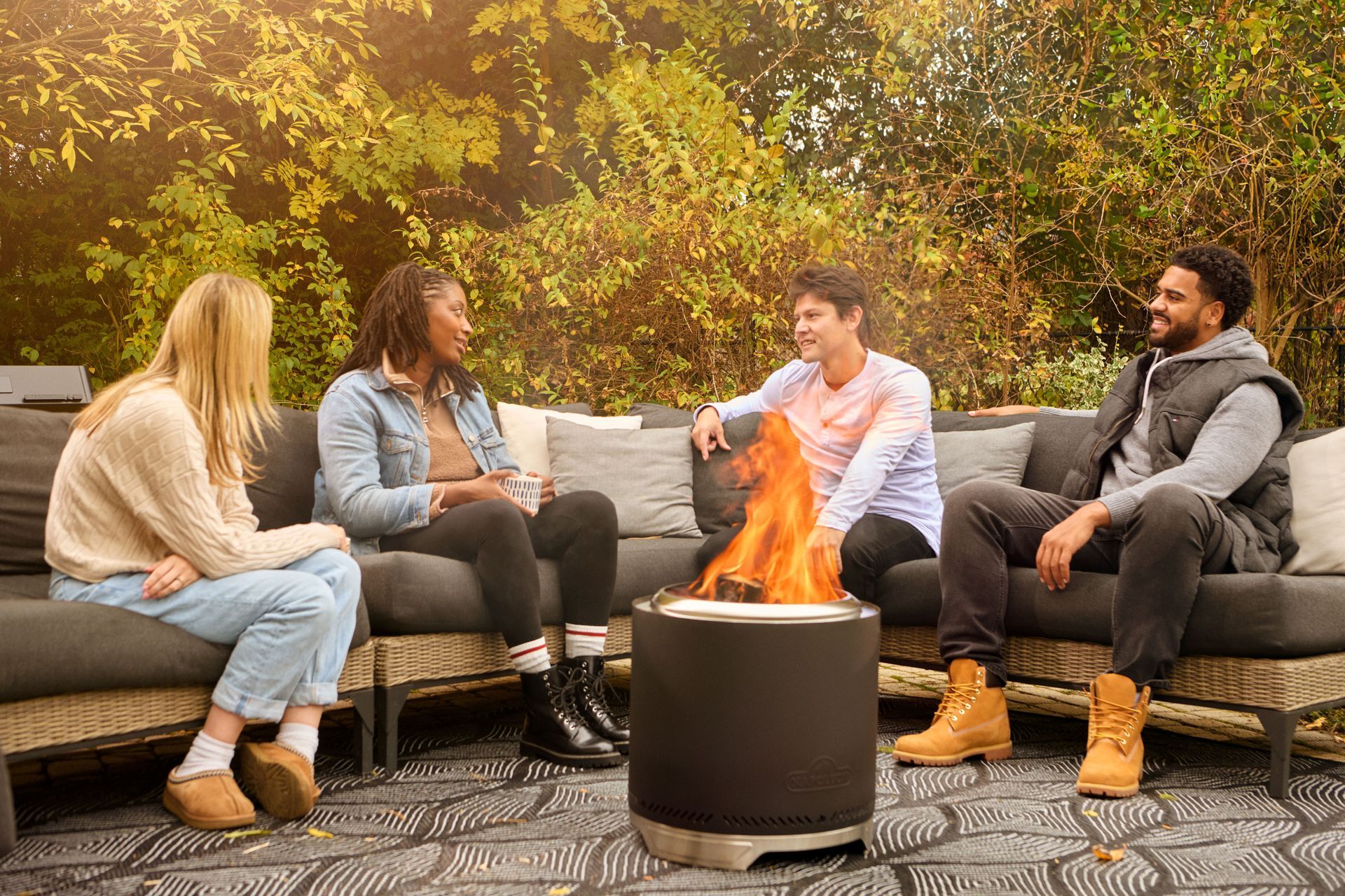 Four people sit on outdoor patio furniture around a circular, lit fire pit in a backyard setting during the daytime.