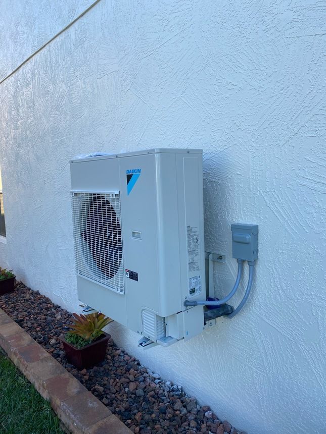 An air conditioning unit mounted on a textured, light-colored wall, with a small electrical box to the right.