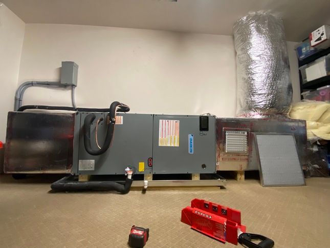 HVAC unit in a room with tools on the floor. The unit is gray, with ductwork and a cylindrical insulated tank.