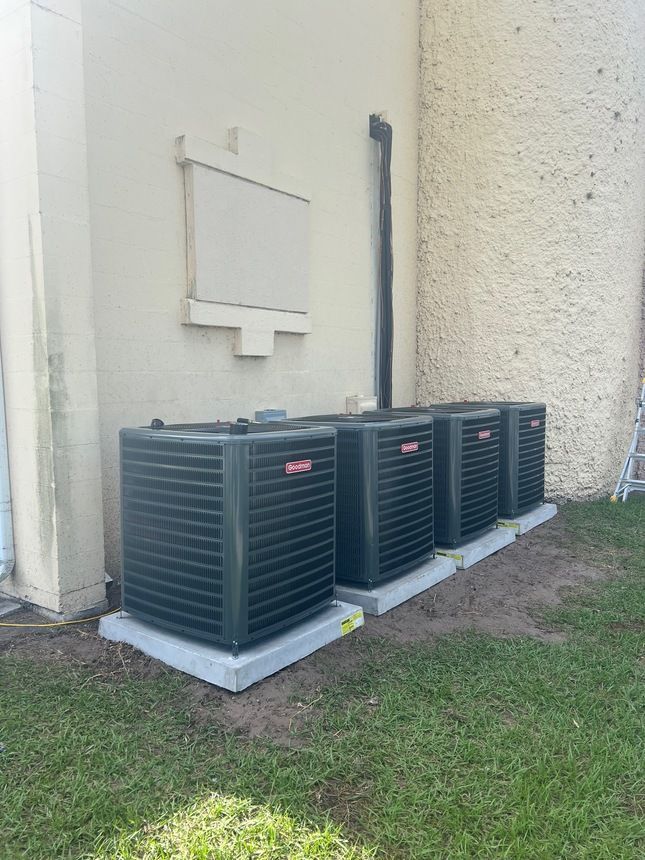 Four air conditioning units on concrete pads against a beige building exterior and green grass.