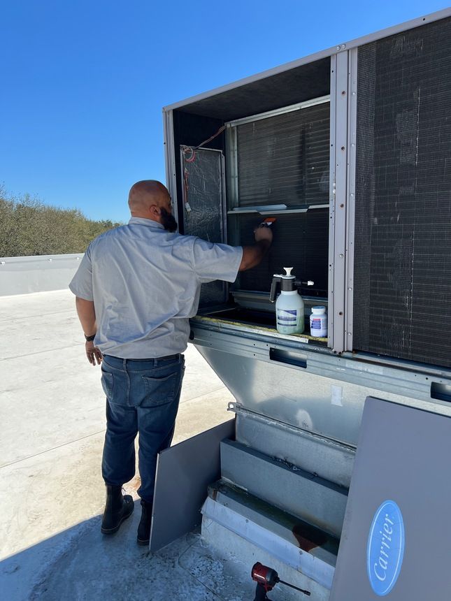 A person cleans an outdoor Carrier HVAC unit on a rooftop. They use a spray bottle and cleaner.