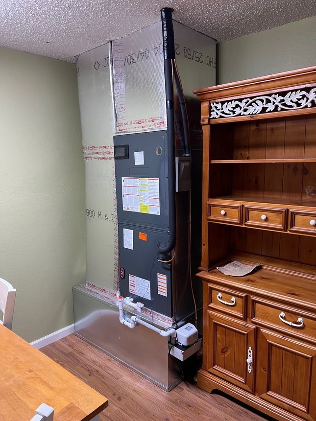 HVAC unit in a room, next to a wooden cabinet. The unit is tall and silver. There's a table and chair to the left.