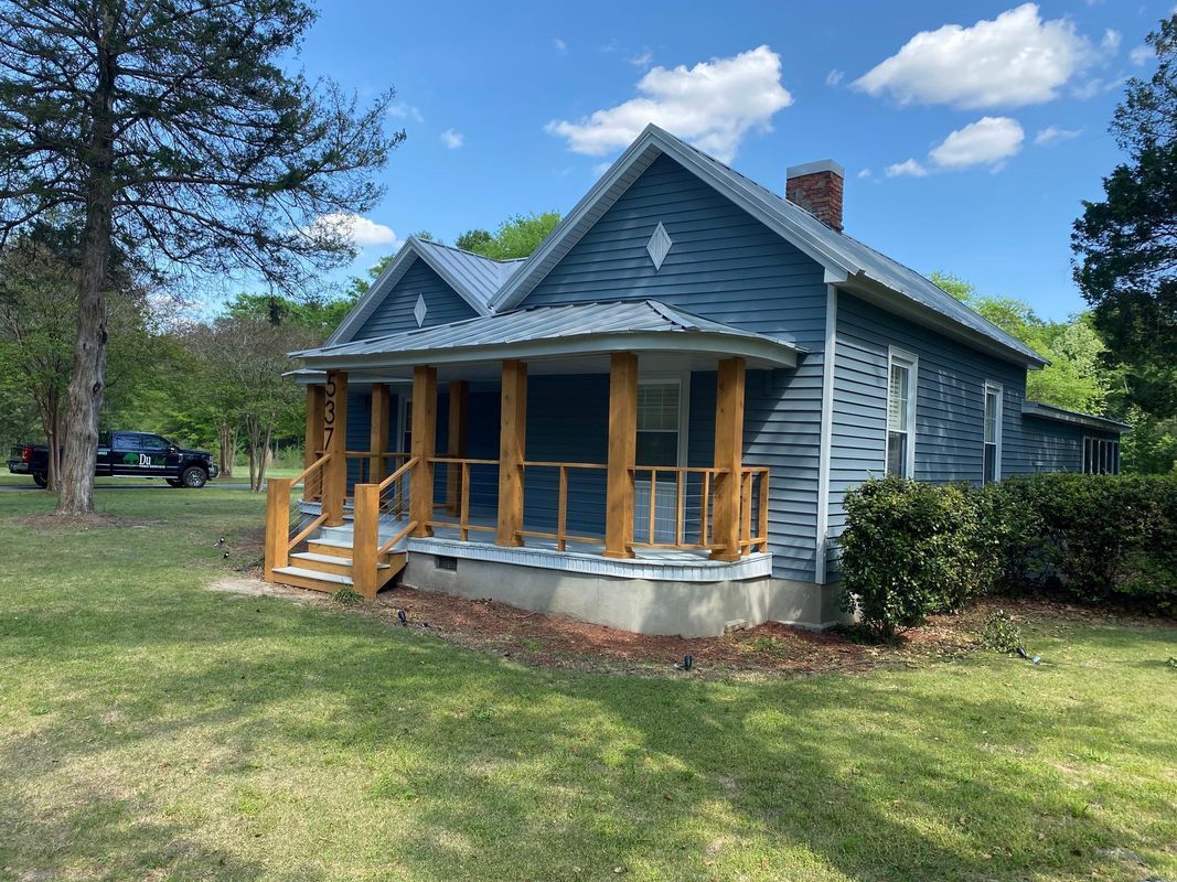 Blue house with front porch, wooden posts, metal roof, green lawn, trees, and a cloudy sky.