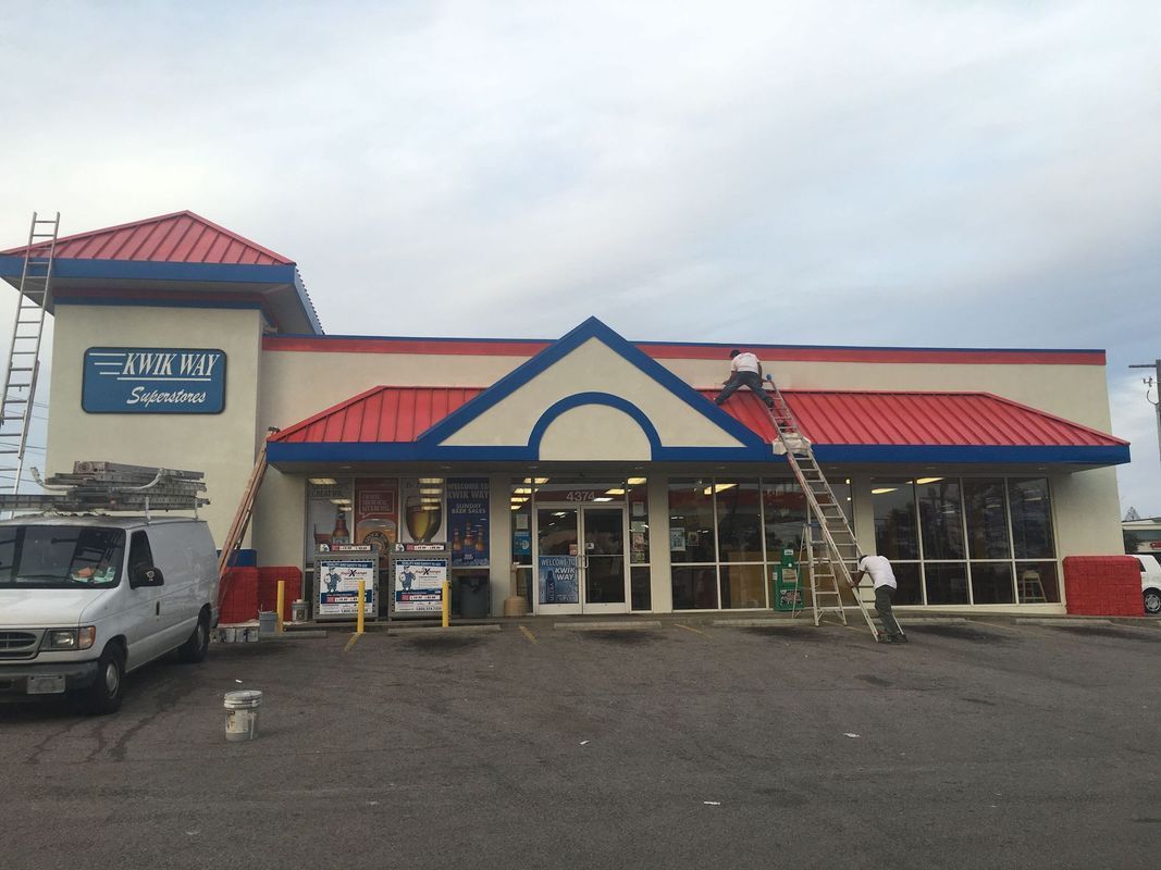 Exterior of a store with red roof, blue trim, and a white van. Workers on ladders, overcast sky.