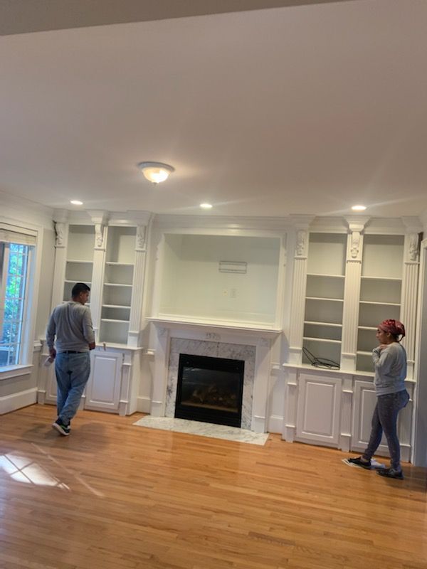 Two people stand in a living room with built-in bookshelves flanking a fireplace; wood floors.