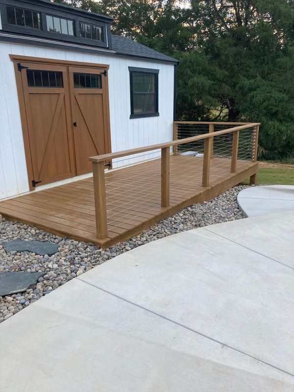 Wooden ramp leading to a white shed with brown doors. Stainless steel railing. Concrete and gravel surround.