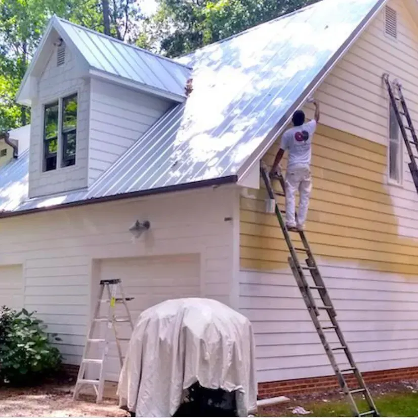 Person painting yellow siding on a white house, standing on a ladder.