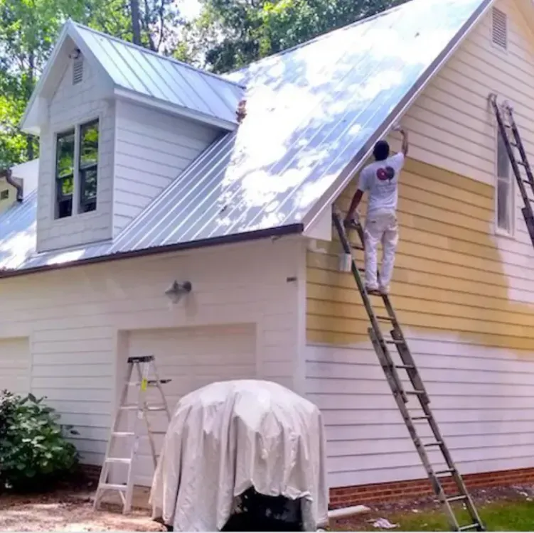 Person painting yellow siding on a white house, standing on a ladder.