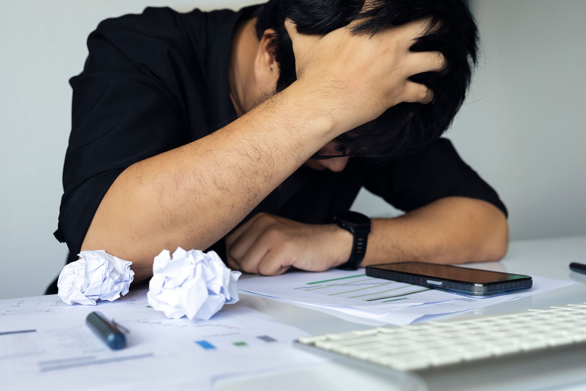 A person slumped over a desk, head in their hands, surrounded by crumpled paper, a smartphone, and business documents.