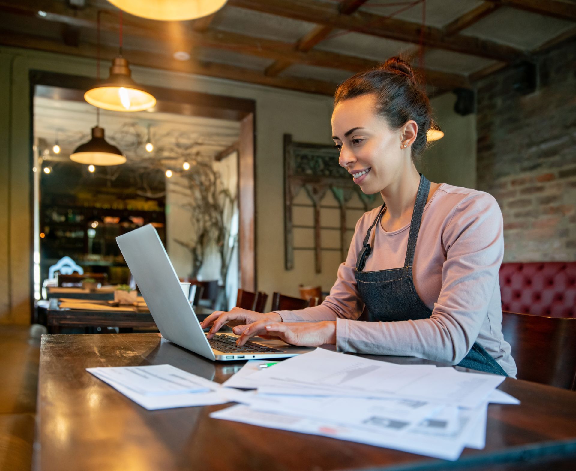 Person in apron working on laptop with papers spread out in cozy café-style setting.